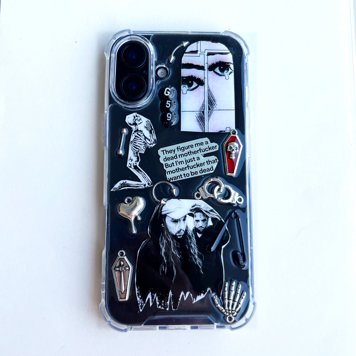 $uicideboy$ g59 phone case featuring a gothic horror-themed design with silver and black charms, including a skeleton, a skull, a vampire illustration, a small coffin, and a quote bubble that reads, 'They figure me a dead motherfucker but I'm just a motherfucker who wants to be dead.'.