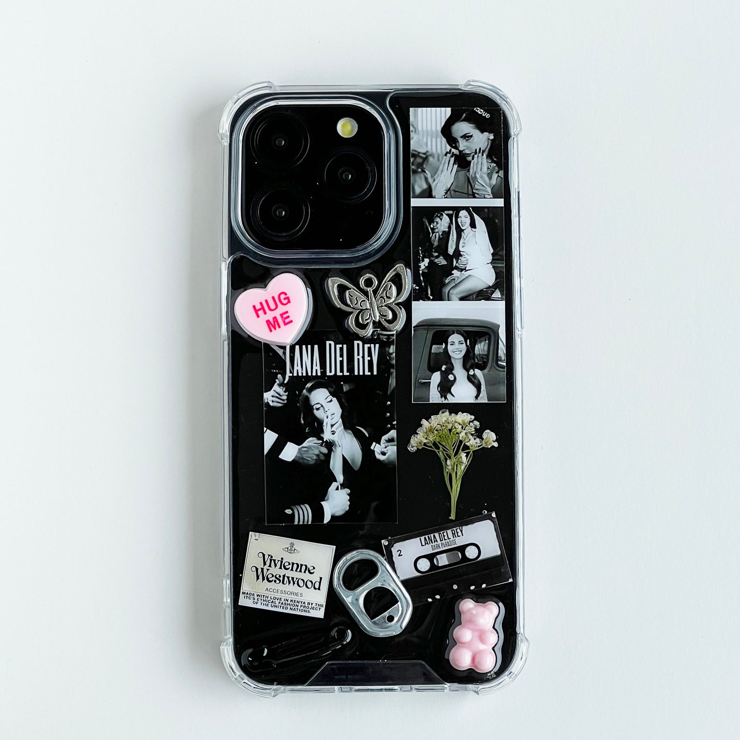 lana del rey collage resin custom phone case featuring a black collage design with photos of Lana Del Rey, a pink heart sticker that reads 'HUG ME', a butterfly charm, cassette tape, bottle opener, and small decorative elements, shown on a light gray background.