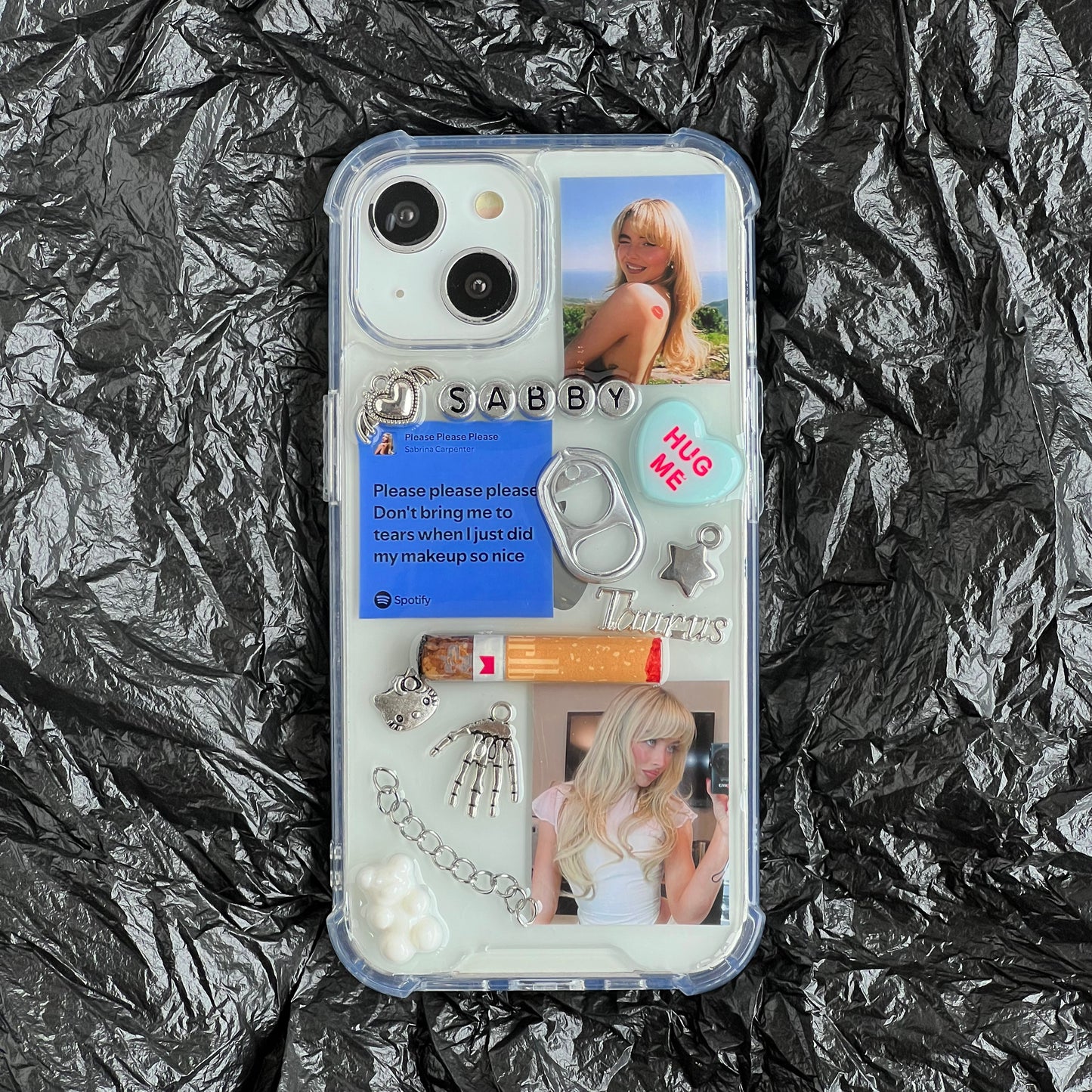 Sabrina Carpenter resin phone case decorated with various pop-culture and fashion-themed charms, including a 'SABBY' nameplate, a blue 'Please please please' card, a silver ring pull, and two photos of a blonde woman, shown flat-laid on a crumpled black background.