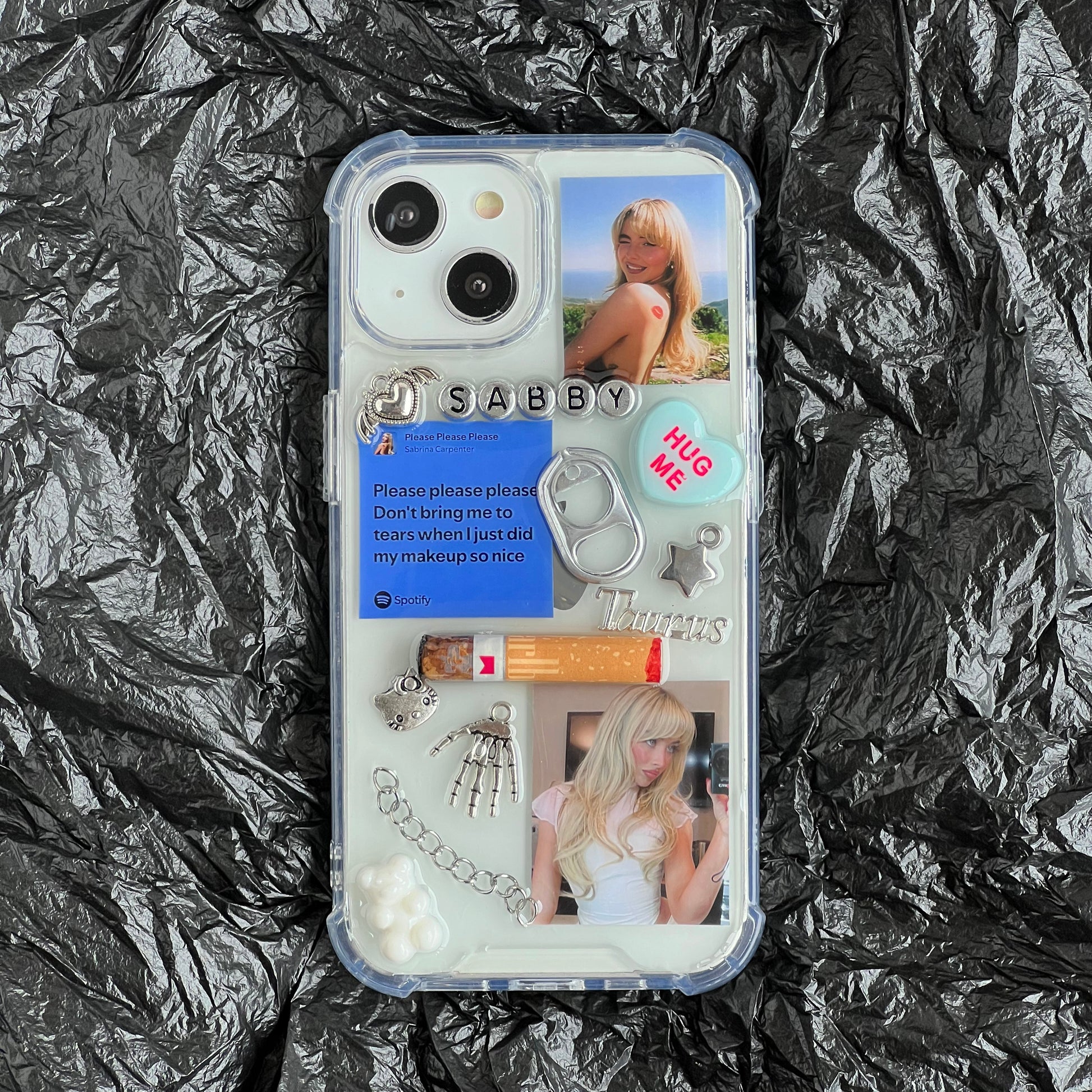 Sabrina Carpenter resin phone case decorated with various pop-culture and fashion-themed charms, including a 'SABBY' nameplate, a blue 'Please please please' card, a silver ring pull, and two photos of a blonde woman, shown flat-laid on a crumpled black background.